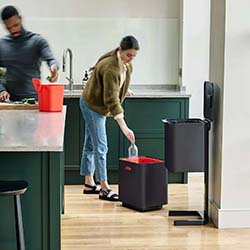 A woman in casual clothing recycles a bottle in a modern kitchen. A man in the background prepares food. The room has a clean and organized atmosphere.