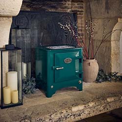 A green wood-burning stove set in a rustic stone fireplace, flanked by a lantern with candles and a vase with branches, creating a cozy ambiance.