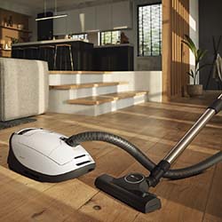 A modern, sunlit living room with wooden floors features a sleek white vacuum cleaner in the foreground. Cozy sofas and a kitchen are in the background.