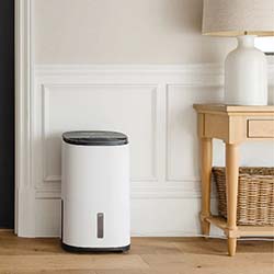 A sleek white air purifier sits on a wooden floor beside a light-colored table with a woven basket. The setting feels clean and minimalistic.