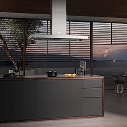 Modern kitchen with a dark island, sleek stovetop, and lemons in a bowl. Large windows reveal a tranquil sunset over the ocean. Minimalist, serene atmosphere.