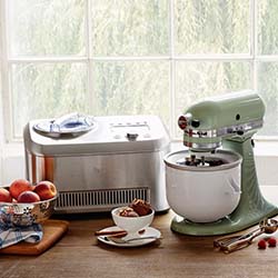 A green stand mixer and a stainless steel ice cream maker sit on a wooden counter. Peaches, ice cream in a bowl, and utensils create a cozy kitchen scene.
