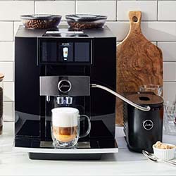 Sleek black espresso machine on a white kitchen counter, with a glass cup of cappuccino and milk frother. Wooden cutting boards in the background. Cozy vibe.