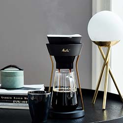 Sleek coffee setup with a modern pour-over coffee maker on a black table, beside a minimalist lamp with a globe shade and stacked books. Cozy atmosphere.