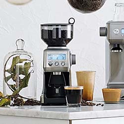Sleek coffee grinder with digital display on a countertop, surrounded by coffee beans, two glass cups, and a plant under a glass dome, evoking a modern cafe vibe.