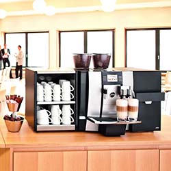Commercial coffee machine on a wooden counter, with shelves holding white mugs. Two lattes are being brewed. Bright room with large windows.