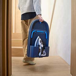 A person carries a compact, folding ironing board with a built-in iron holder up wooden stairs. The setting is modern and minimalistic.