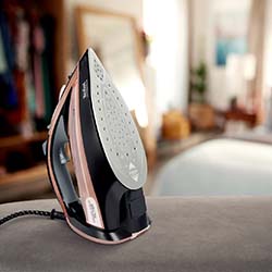 A modern steam iron with a black and rose gold design rests on an ironing board in a cozy, softly-lit bedroom. The room is slightly blurred in the background.