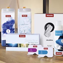 A collection of Miele laundry products on a wooden surface. Includes detergent bottles, wipes, and detergent pods, conveying a clean, organized feel.