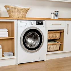 A modern white washer in a laundry room, surrounded by wooden shelves with towels, wicker baskets, and detergent, creating a tidy, organized space.