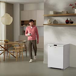 A man in a pink sweater and brown pants walks in a modern kitchen, holding a cup. The room features light wood furniture and a white appliance.