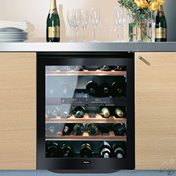 A wine cooler with glass door, showcasing several bottles on wooden shelves. Champagne, glasses, and flowers are on the counter, evoking a celebratory feel.