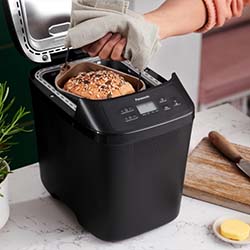 A person uses an oven mitt to remove fresh multigrain bread from a black bread machine on a kitchen counter, near a cutting board and butter.