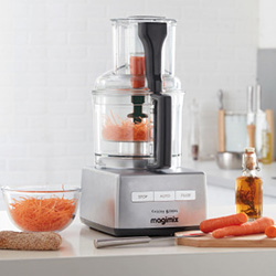 A modern food processor with shredded carrots inside sits on a kitchen counter. Nearby are whole carrots, a glass bowl of shredded carrots, and a small bottle of oil. The setting is bright and tidy.