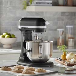 A black stand mixer is on a kitchen counter, surrounded by freshly baked cookies, fruit, and spices. The scene conveys a warm, inviting baking atmosphere.