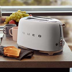White Smeg toaster on a kitchen counter with toasted bread, surrounded by cups and fresh fruit, creating a cozy morning atmosphere.