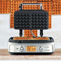 A stainless steel waffle maker with an open lid displays golden waffles inside. A digital timer reads 3:29. Background shows a waffle wall.