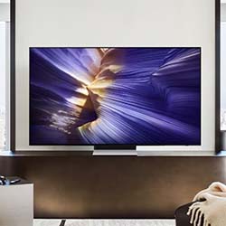 A modern living room features a large, wall-mounted TV displaying vibrant abstract art in purple and gold tones. A cozy blanket is draped over a chair nearby.