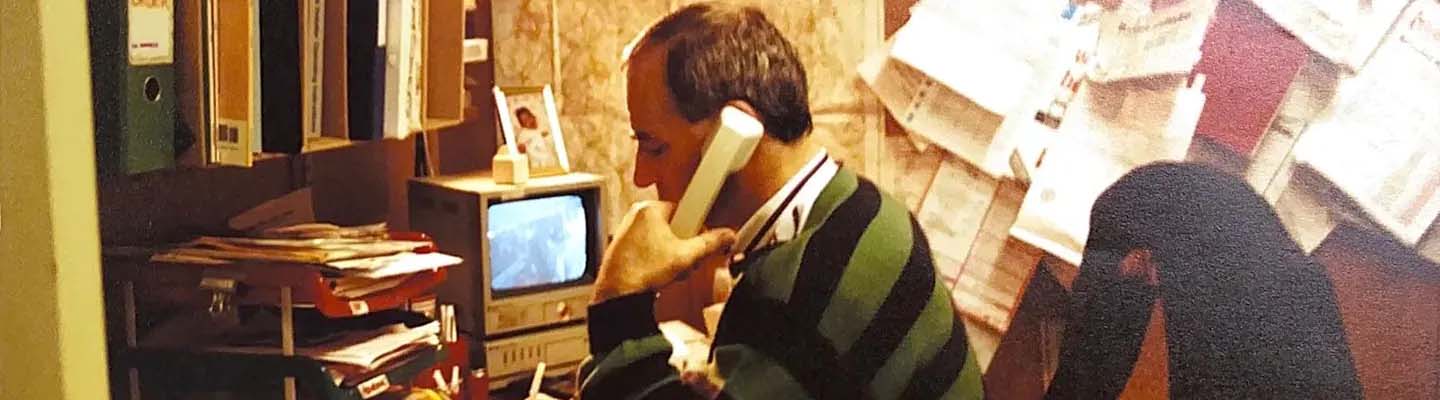A man in a green striped sweater is on a landline phone in a cluttered office, with binders, a stack of papers, a small TV, and a wall of notes. The scene conveys a busy, retro work environment.