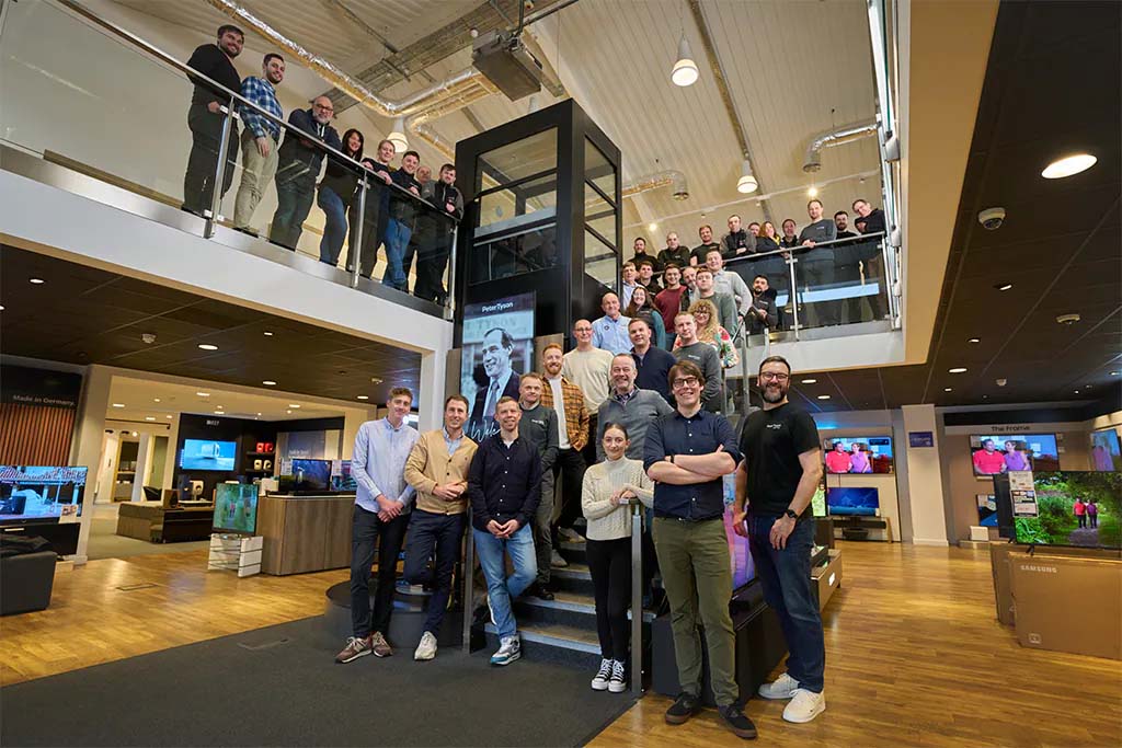 A large group of people pose inside a modern store. Some are standing on a staircase while others are on the balcony above, smiling in a lively, welcoming atmosphere.