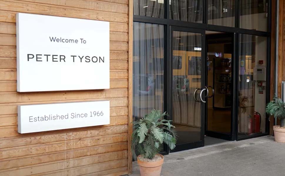 Entrance of a building with wooden siding, displaying signs that read 'Welcome to Peter Tyson' and 'Established Since 1966.' Flanked by potted plants.