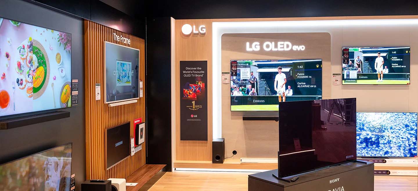 Display room showcasing various TVs on a wooden wall, including LG OLED models. Screens show vibrant content, creating a modern, tech-savvy atmosphere.