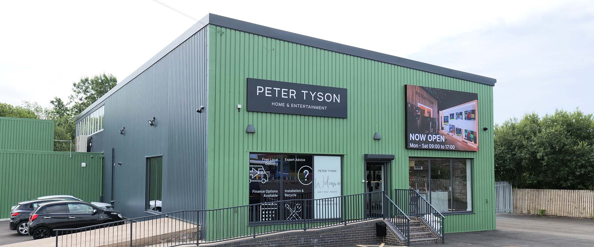 A modern, green industrial building with large signs for "Peter Tyson Home & Entertainment." The exterior features windows and an entrance ramp. A billboard announces "Now Open" with operating hours. Three parked cars are visible on the left, and there are trees in the background, suggesting a welcoming and professional environment.