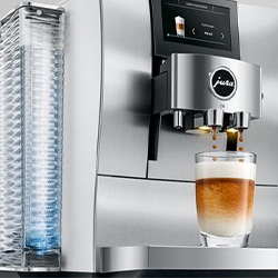 Sleek silver espresso machine pouring coffee into a clear glass, which has layers of milk and espresso. A blue backlit water tank is visible on the side.