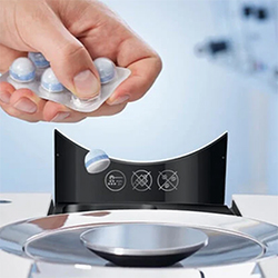 A hand places a detergent pod into a modern washing machine dispenser. The background is blurred, emphasizing the pod's vibrant colors and sleek design.