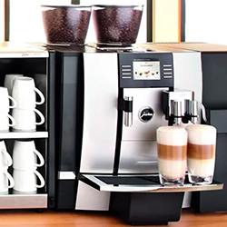 A sleek espresso machine on a counter with dual bean containers, two lattes in glass cups, and a shelf holding stacked white mugs, conveying a modern, café vibe.