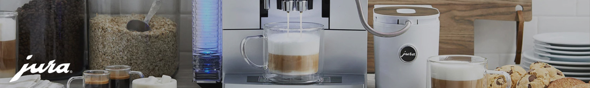 Coffee station with espresso machine preparing a cappuccino in a glass mug. Nearby are jars of coffee beans, an oat container, and a plate of cookies.
