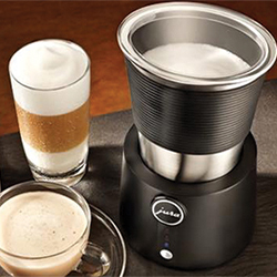 A black milk frother with foamy milk beside two cups of coffee, one with layered foam. Warm, inviting setting with a focus on coffee preparation.