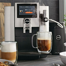 A sleek espresso machine on a kitchen counter brews coffee into a glass mug. Foam-topped cappuccino sits beside it. The scene feels modern and inviting.