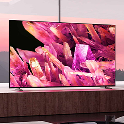 A flat-screen TV displays vivid pink and purple crystals, creating a vibrant and dynamic visual. The TV sits on a wooden surface against a sunset backdrop.