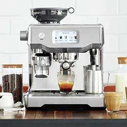 A sleek, silver espresso machine on a wooden counter, brewing coffee into a clear glass. Surrounding are jars of beans, milk, and a silver frothing pitcher, against a white tile backdrop.