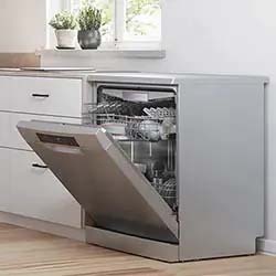 Open stainless steel dishwasher in a modern kitchen, door ajar revealing racks inside, against a light wood floor with a plant on the counter.