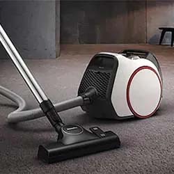 A modern canister vacuum cleaner with a sleek white and black design is on a carpeted floor in a minimalist room, conveying efficiency and style.