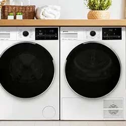 Front view of a modern washer and dryer set with digital displays, set under a wooden countertop. Towels and a potted plant add a homely touch.