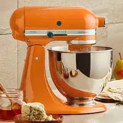 Orange stand mixer with a shiny stainless steel bowl sits on a kitchen countertop. Nearby are jars of honey and sliced bread, creating a warm, homey feel.