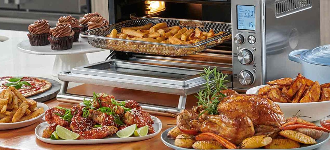 A countertop oven with fries inside sits amid a spread of roasted chicken, spicy wings with lime, a mini pizza, cupcakes, and seasoned fries.
