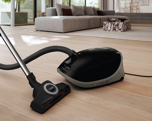 A sleek black vacuum cleaner with a hose and pole attachment is on a light wooden floor. The cord trails behind, creating a tidy, efficient atmosphere.