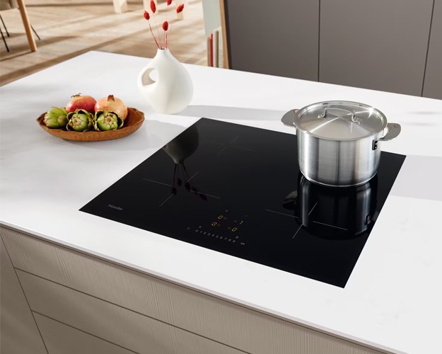 A modern kitchen with a sleek black induction cooktop and a stainless steel pot. Nearby, a wooden bowl holds artichokes and apples, creating a clean, inviting atmosphere.