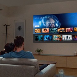 A couple sits on a sofa in a cosy living room, watching a large screen displaying a streaming platform with various movie thumbnails and a sci-fi theme.