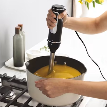 A person uses an immersion blender to puree soup in a pot on a stove. Two bottles and a small bowl are in the background, lit by natural light.