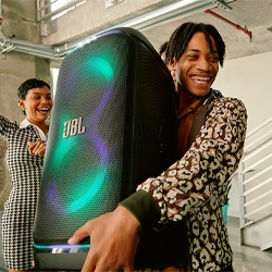 A joyful young man in a patterned shirt holds a large JBL speaker with colourful lights. A woman behind him is clapping, set in an industrial space.
