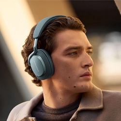 Young man with short curly hair wearing large over-ear headphones, looking to the right. He wears a brown jacket, with a blurred urban background.