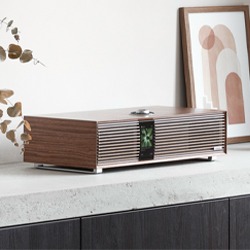 A sleek wooden speaker with a modern design sits on a concrete countertop. Nearby is abstract wall art and dried leaves, creating a minimalist, calm atmosphere.