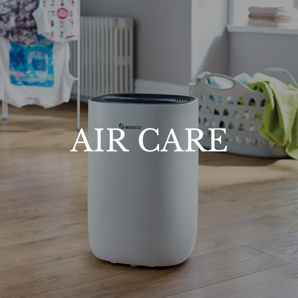 A white air purifier labeled "Meaco" on a wooden floor in a laundry room. Background includes a drying rack and laundry basket. Text reads "Air Care."