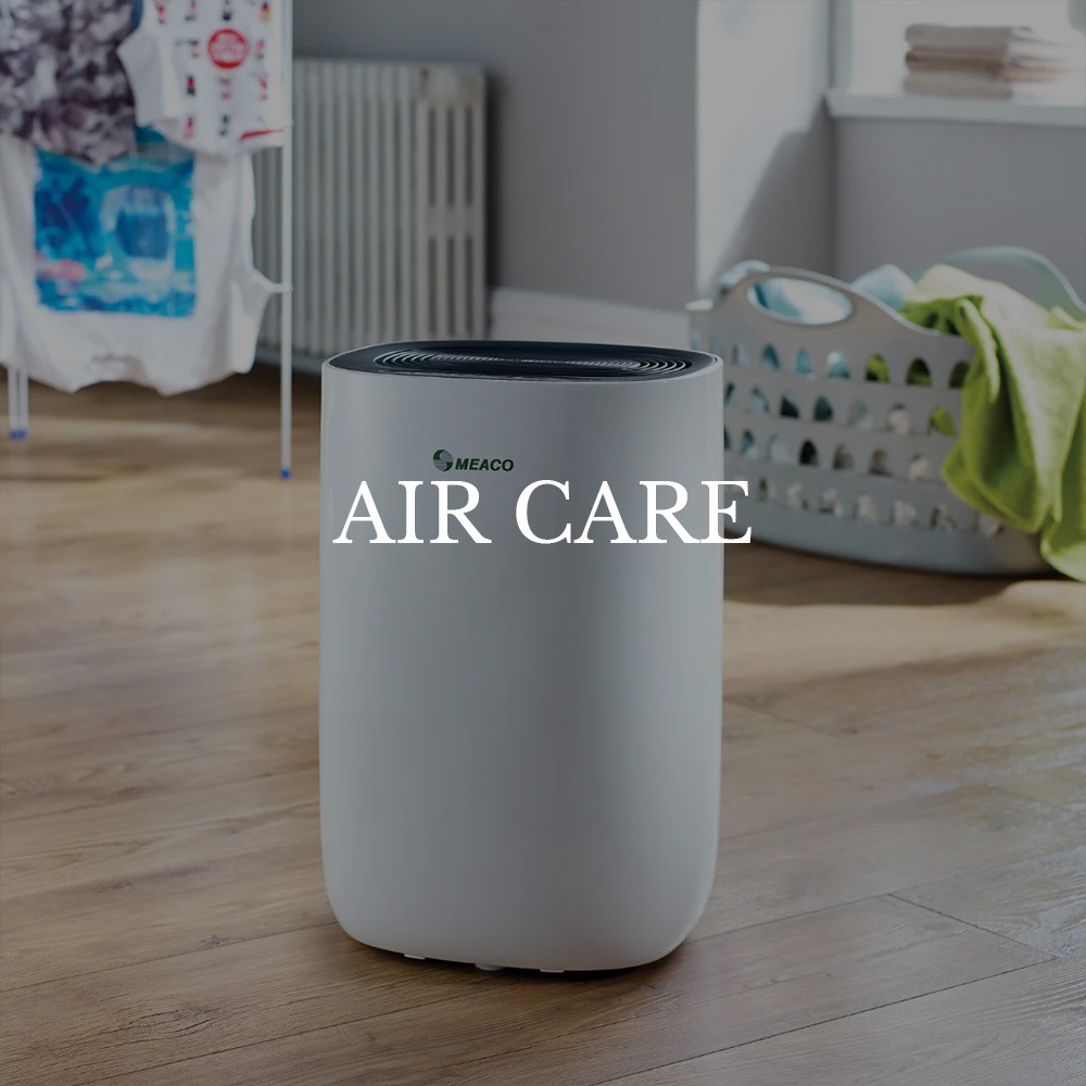 A white air purifier labeled "Meaco" on a wooden floor in a laundry room. Background includes a drying rack and laundry basket. Text reads "Air Care."