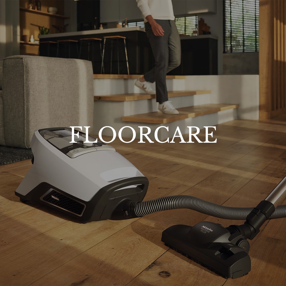 Vacuum cleaner on a wooden floor in a modern living room. A person walks in the background. Text "FLOORCARE" conveys cleanliness and comfort.
